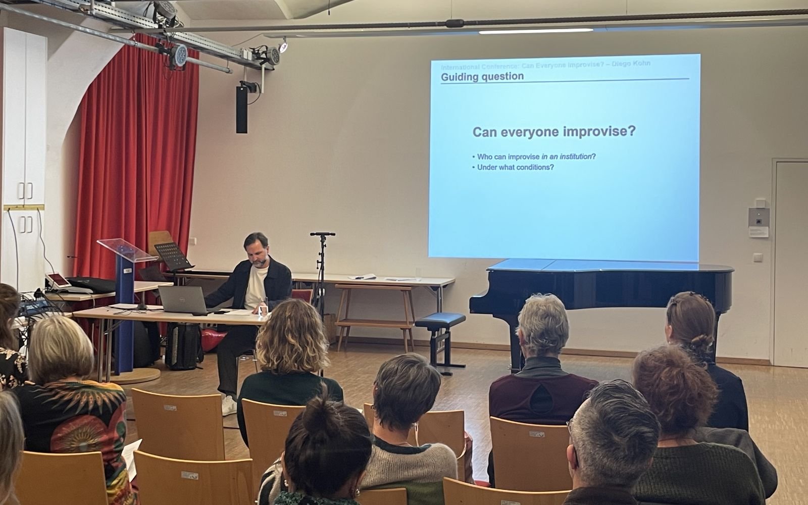 Diego Kohn presenting at a table; the projected slide asks: Can everyone improvise? Who can improvise in an institution? Under what conditions?